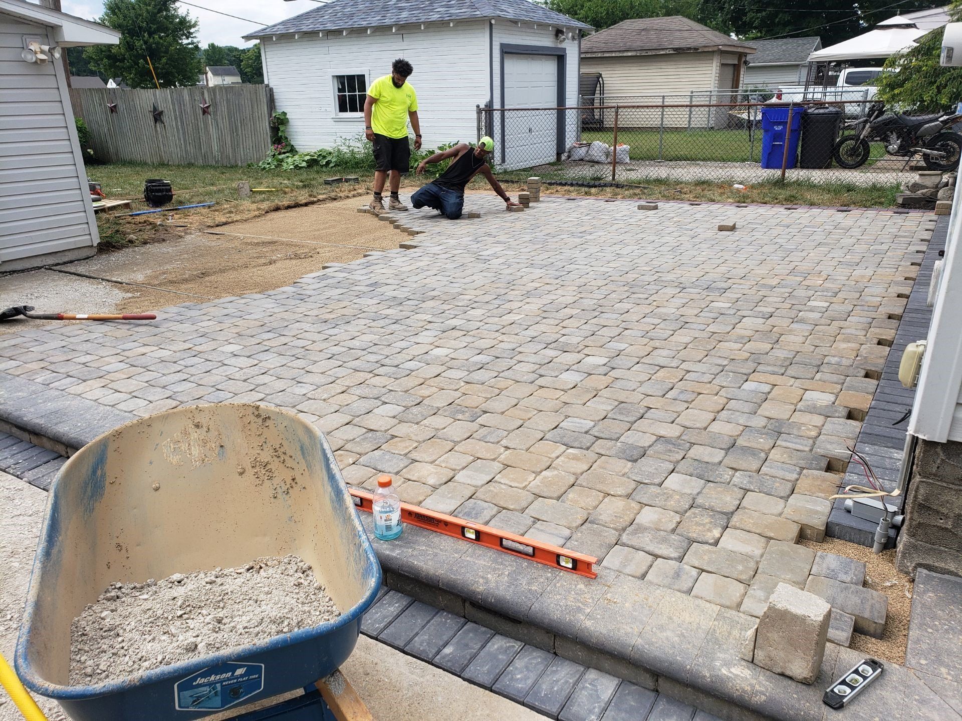 Workers paving a driveway with gray pavers beside a wheelbarrow and level in a residential yard