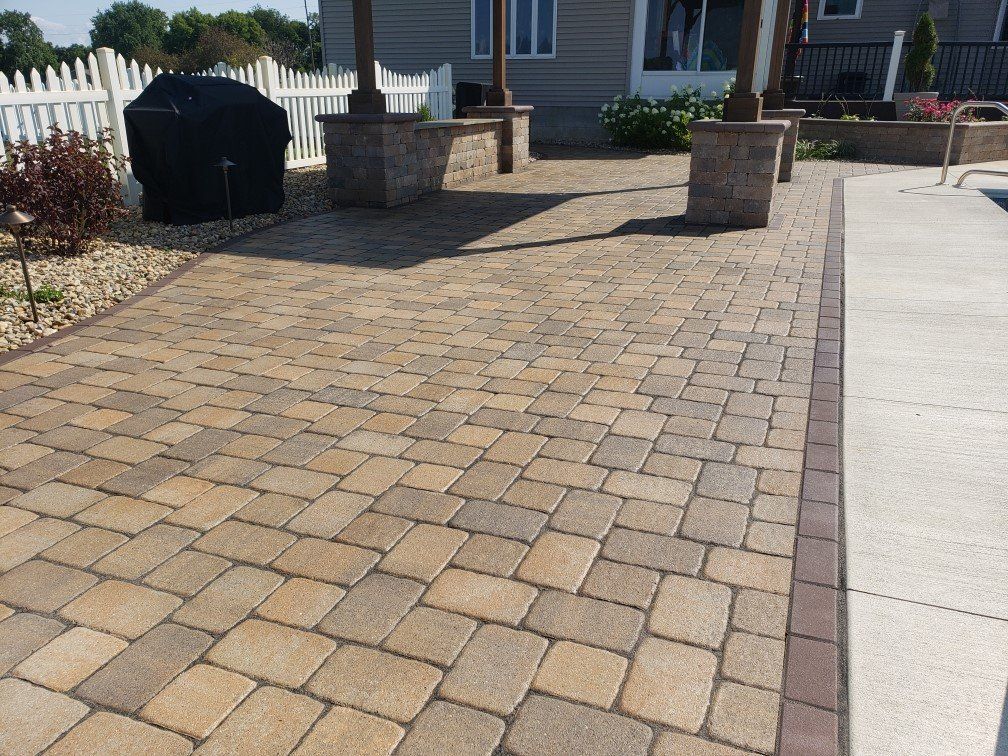 Brick paver walkway leading to a house entrance beside a concrete driveway and stone columns.