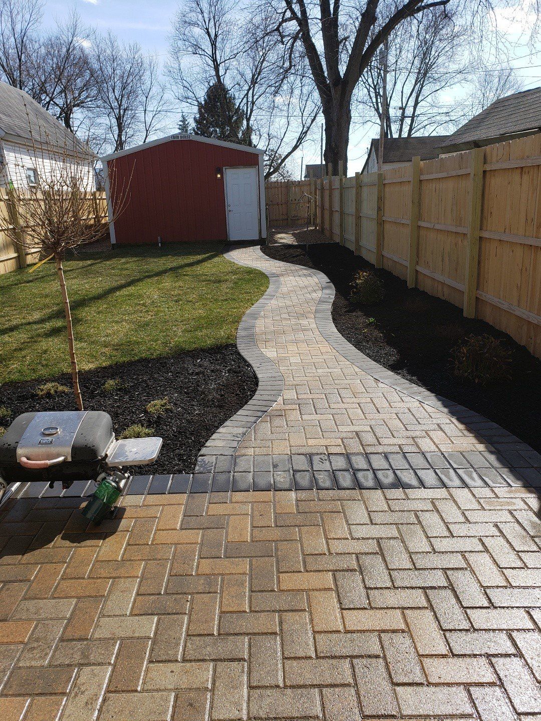 Backyard with brick paver path leading to a red shed, bordered by lawn and a wooden fence.