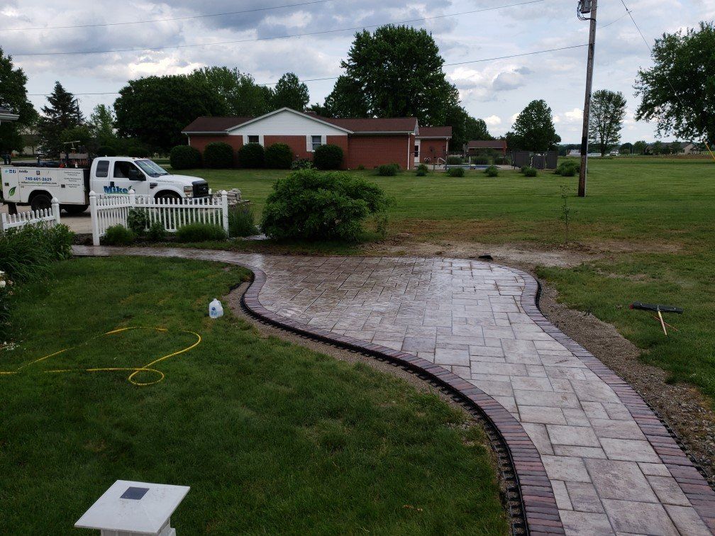 Curved brick walkway through a grassy yard toward a brick building under cloudy skies
