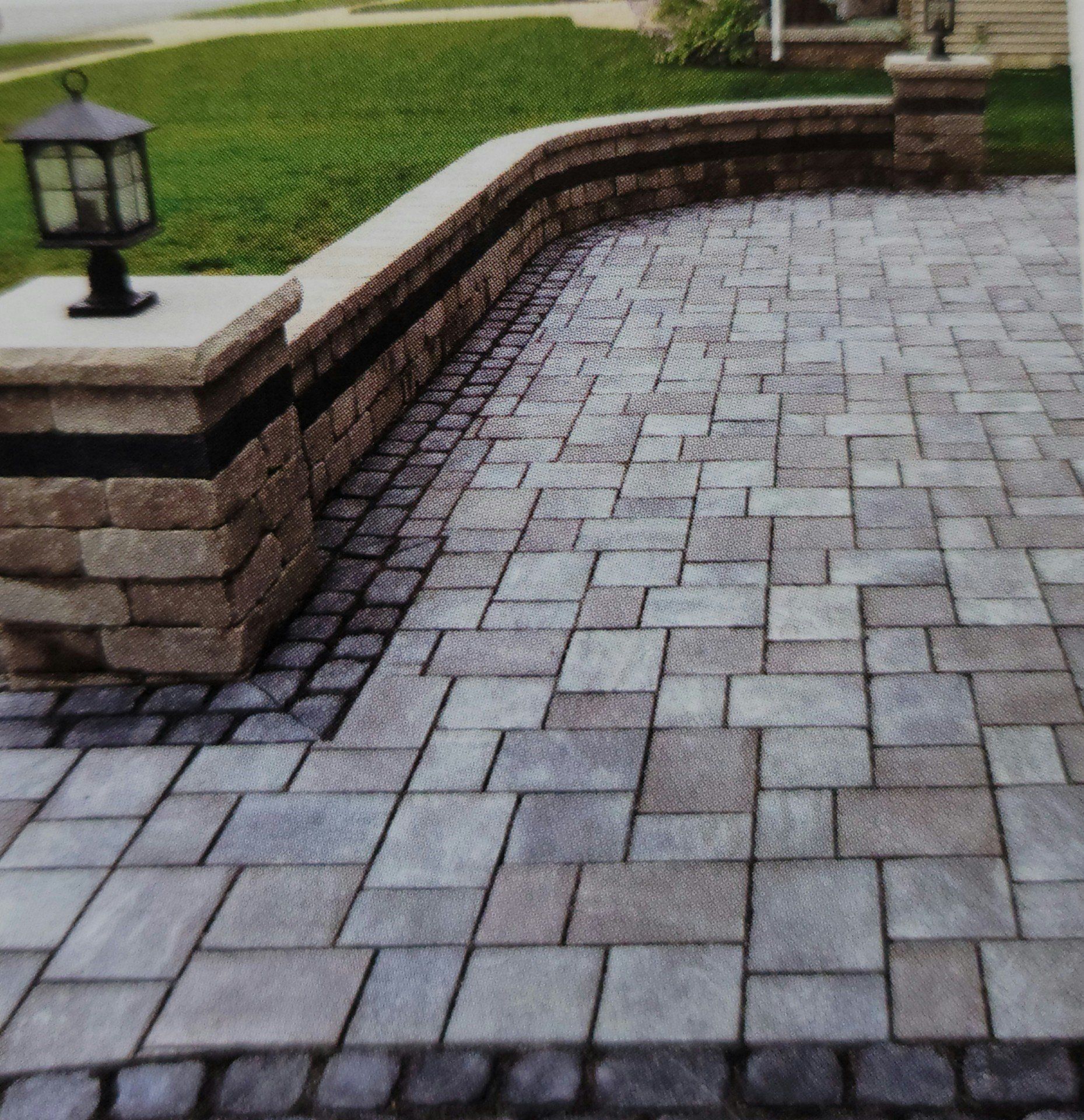 Curved brick paver patio beside a stone retaining wall and lawn with a lantern light.