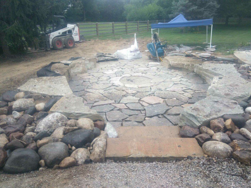 Stone patio under construction with a central fountain, rocks, a blue canopy, and a white tractor in the background