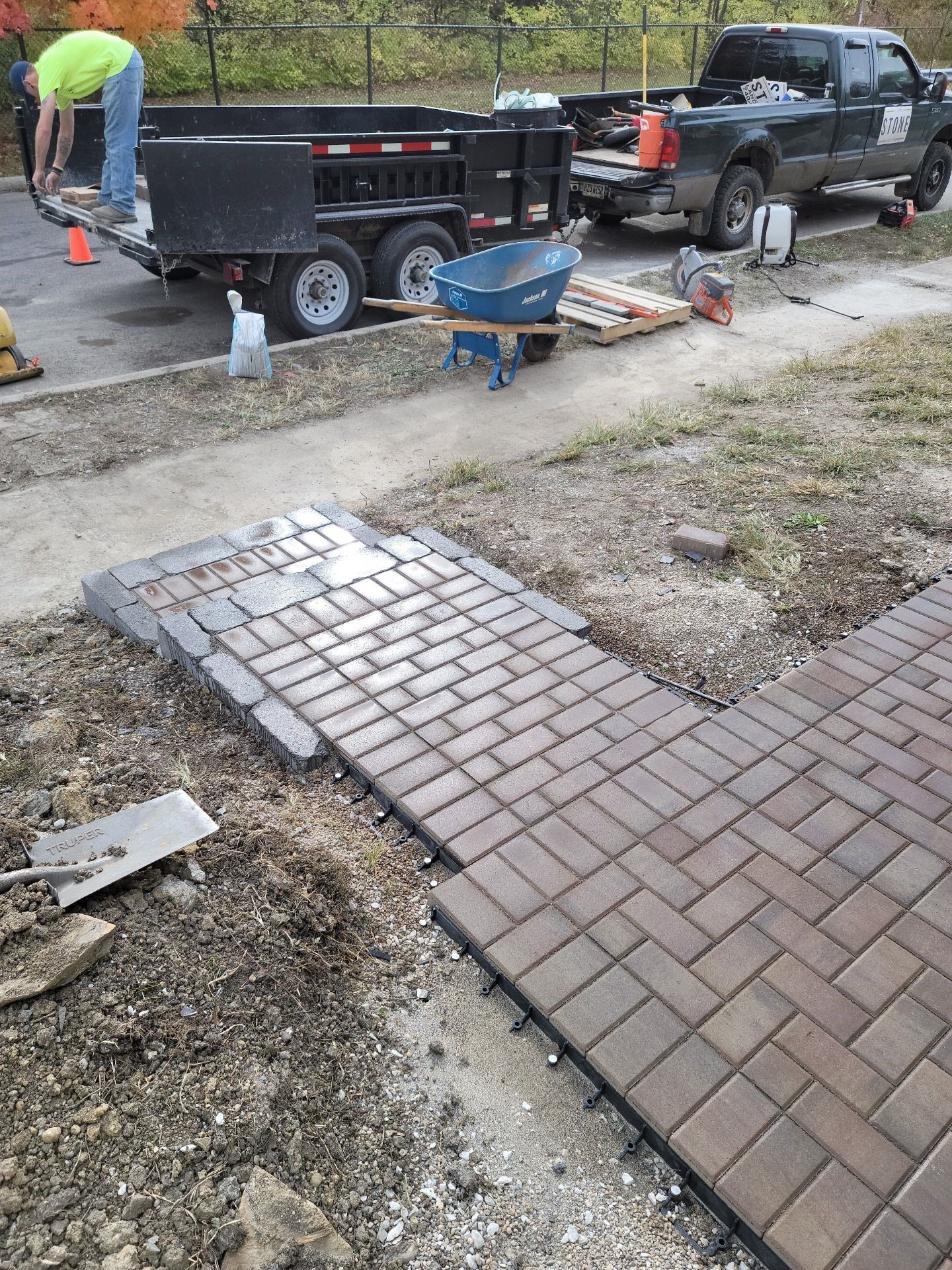 Workers repairing a brick walkway beside parked trucks and tools on a roadside.