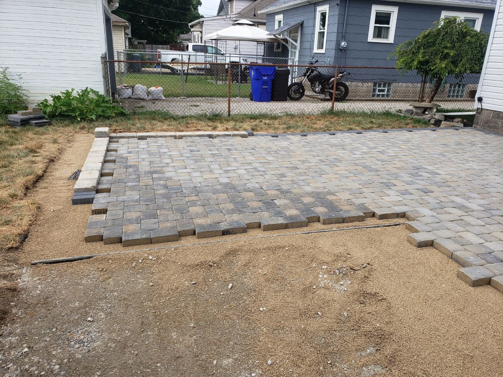 Partially paved backyard patio with gray pavers, bordered by gravel and soil beside a house