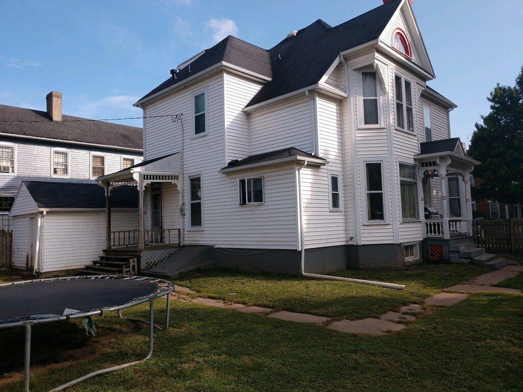White two-story house with black roof and porch, viewed from the yard on a sunny day