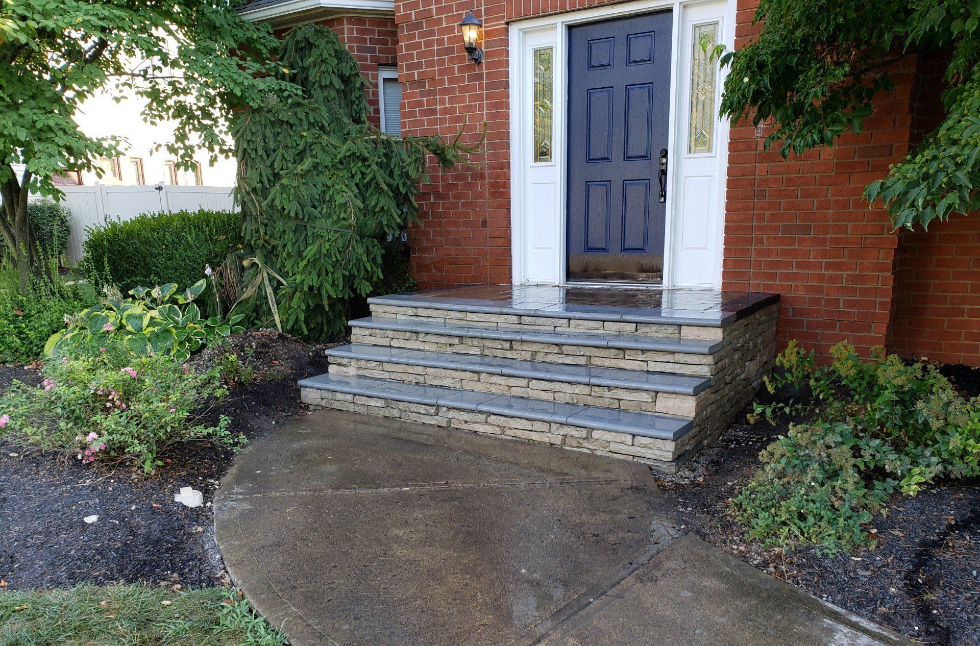 Brick house front entrance with blue door, stone steps, and wet walkway surrounded by shrubs