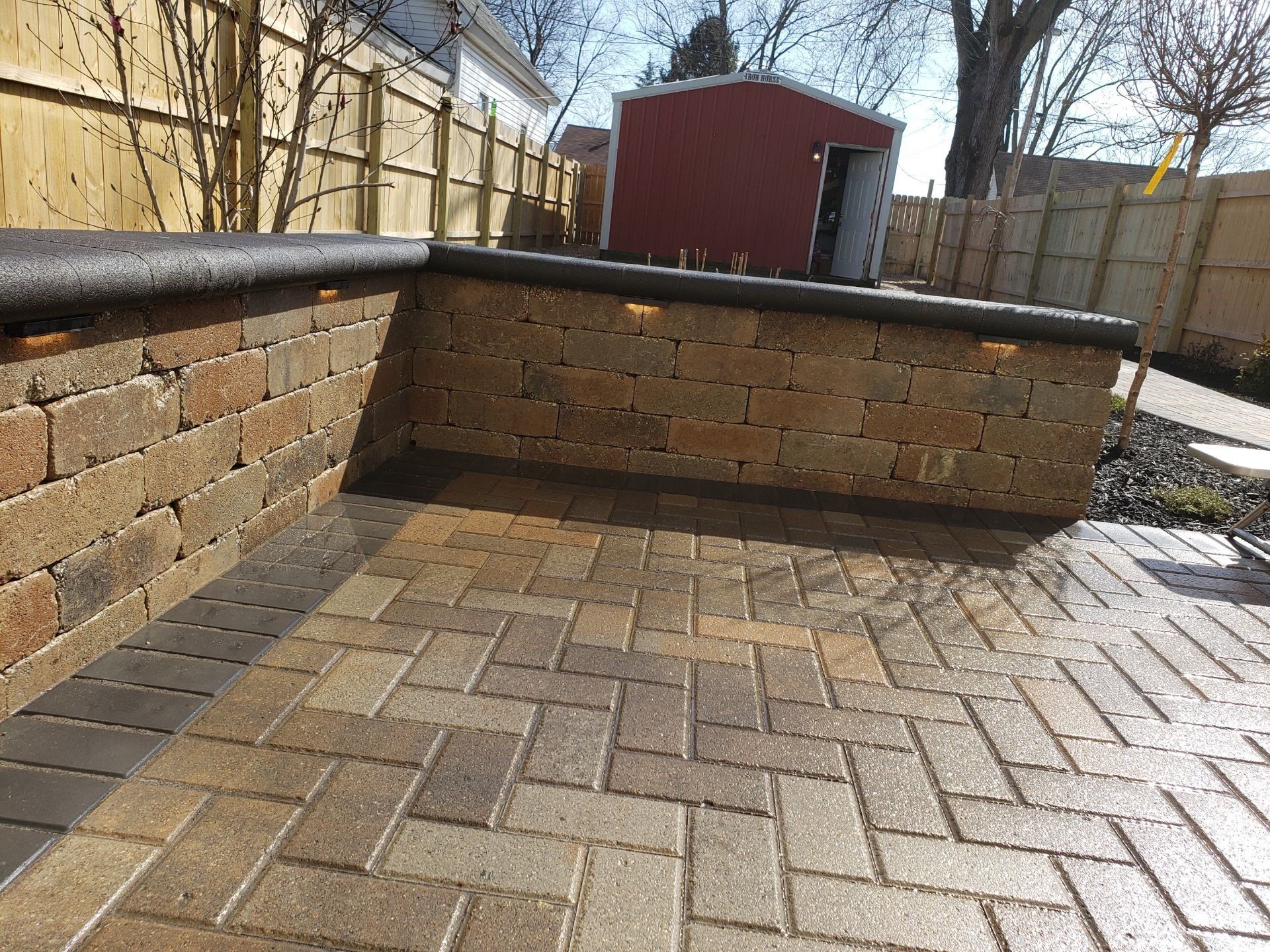 Brick patio with a low curved brick wall and red shed in a fenced backyard