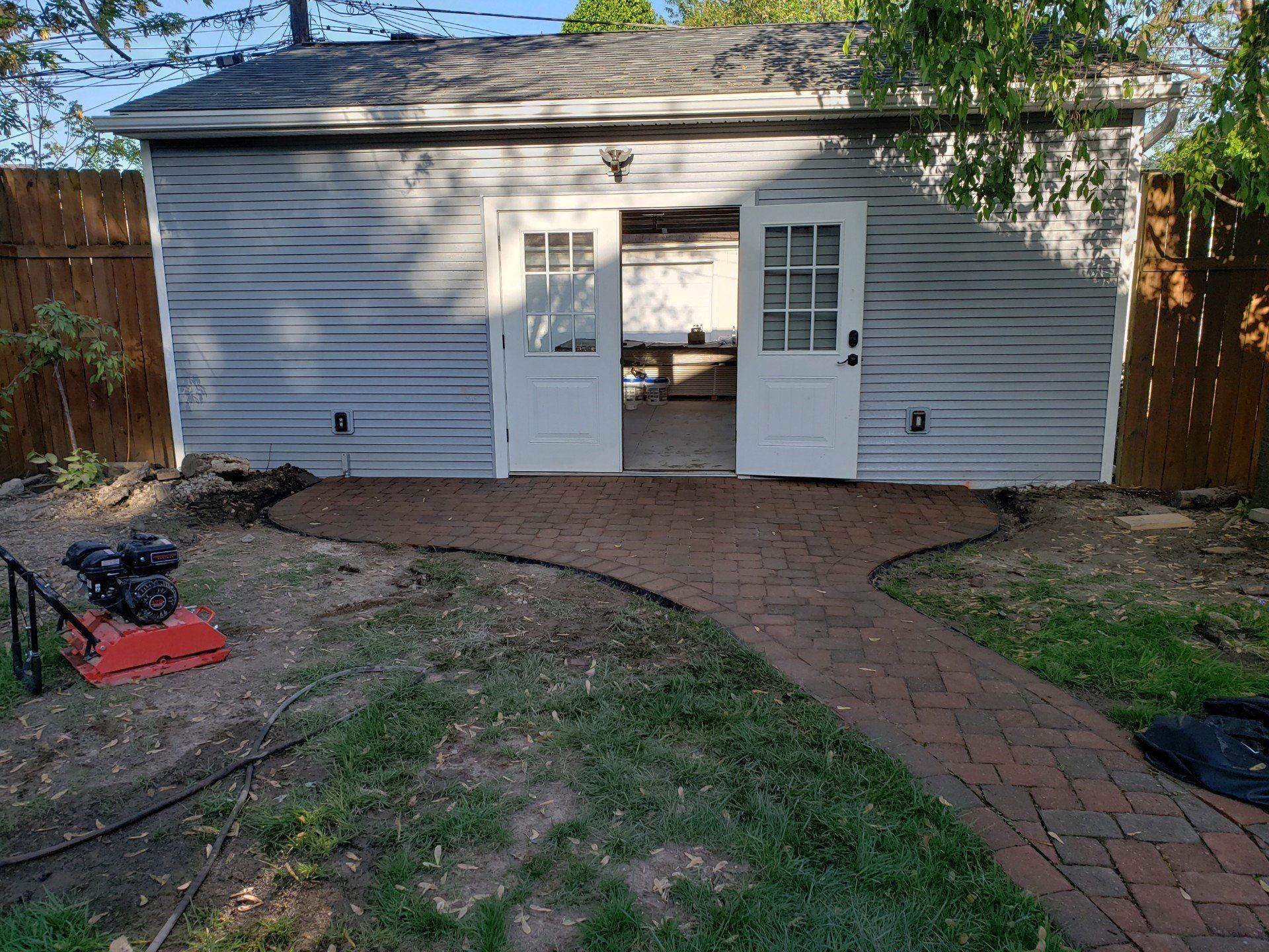 White shed with double open doors and brick walkway in a backyard, with trees and a wooden fence nearby