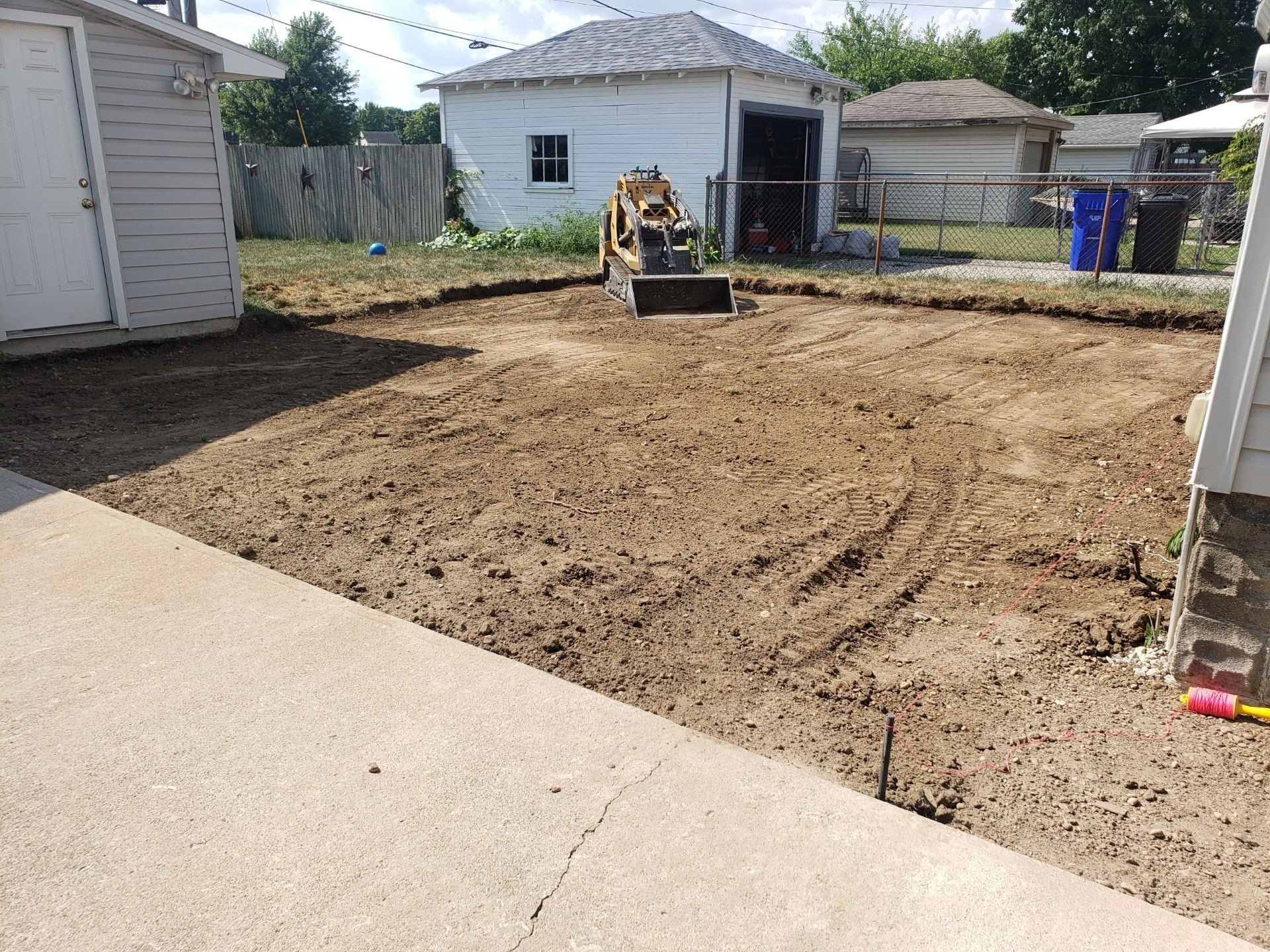 Backyard with freshly graded dirt and a small skid steer near a white shed and fence