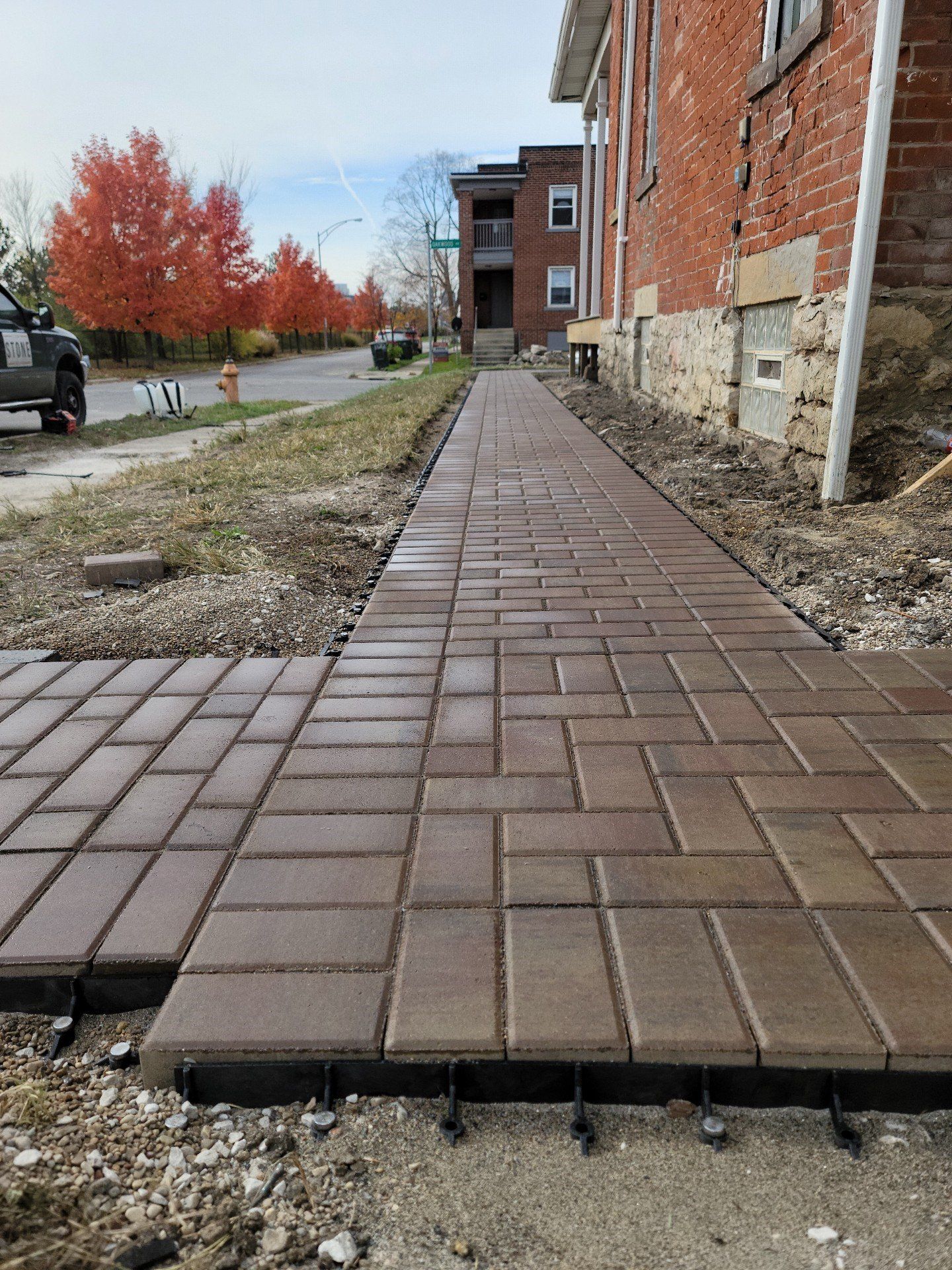 Brick sidewalk beside a brick building, with a damaged edge and autumn trees in the background
