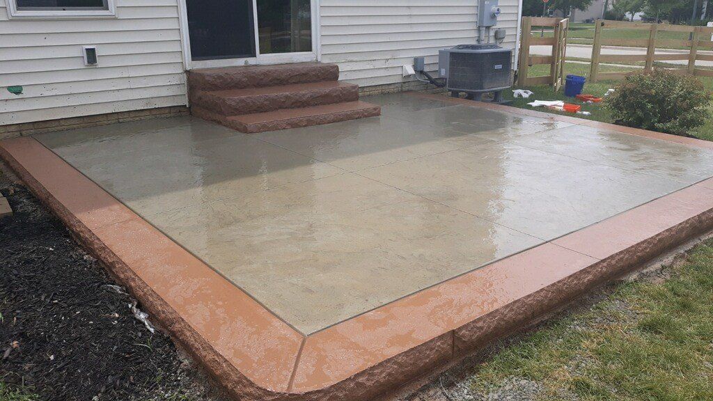 Freshly poured concrete patio with brick border beside a house, wet from rain