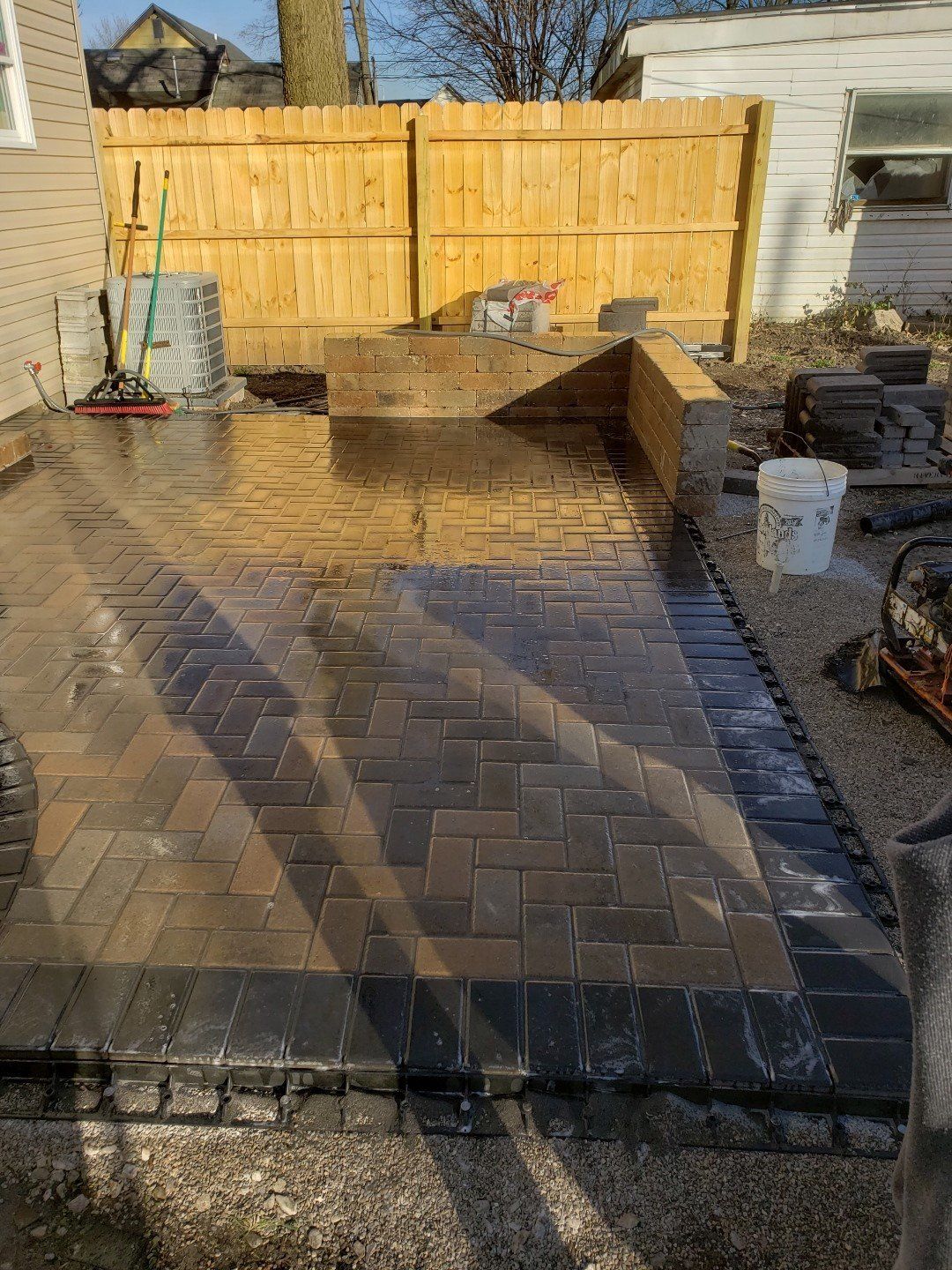 Backyard patio under construction with wet pavers, a wooden fence, and work tools nearby