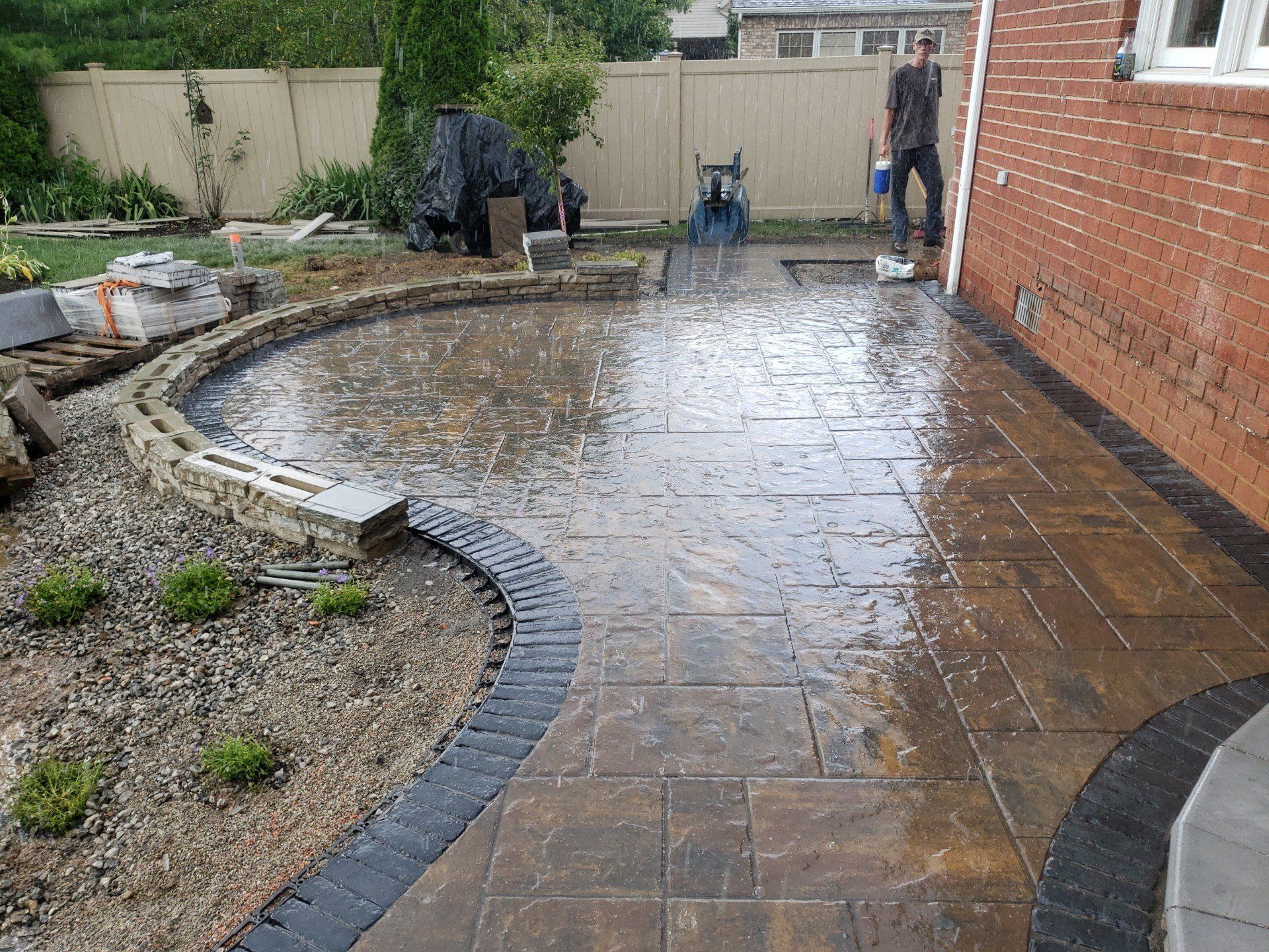 Curved wet stone patio beside a brick wall and gravel garden, with people and tools in the background