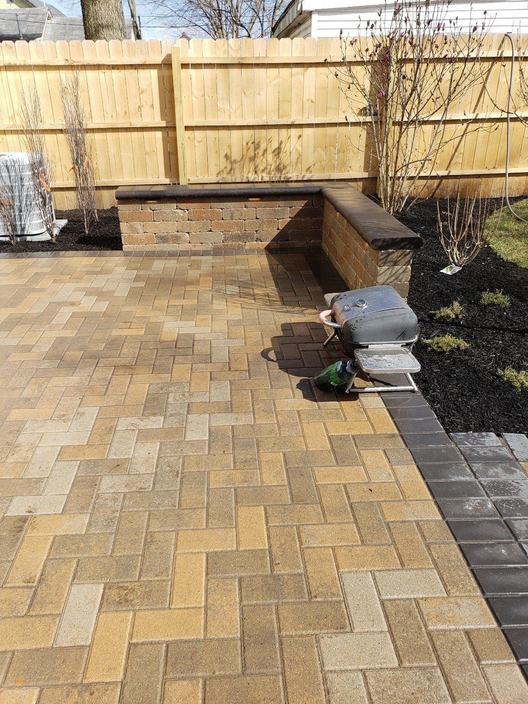 Backyard patio with tan pavers, wooden fence, bench, and a covered grill beside a garden bed