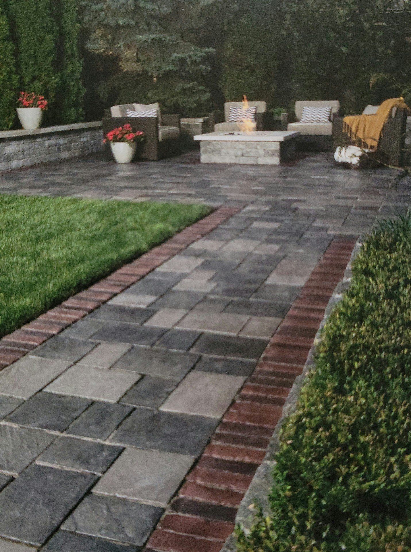 Brick and gray stone garden walkway beside a green lawn and hedges, leading to a patio with chairs and planters