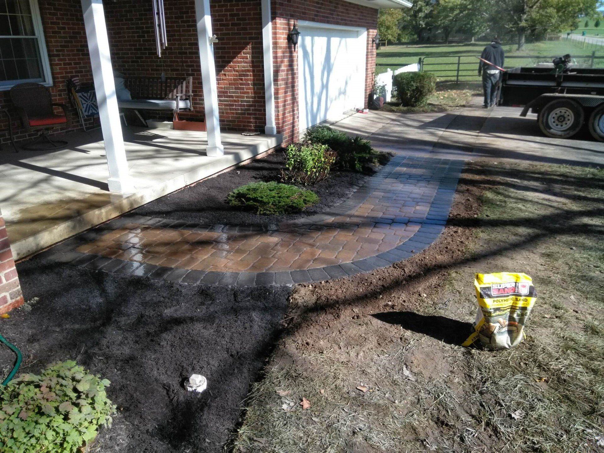Freshly mulched garden beds border a curved brick walkway beside a house porch and driveway.
