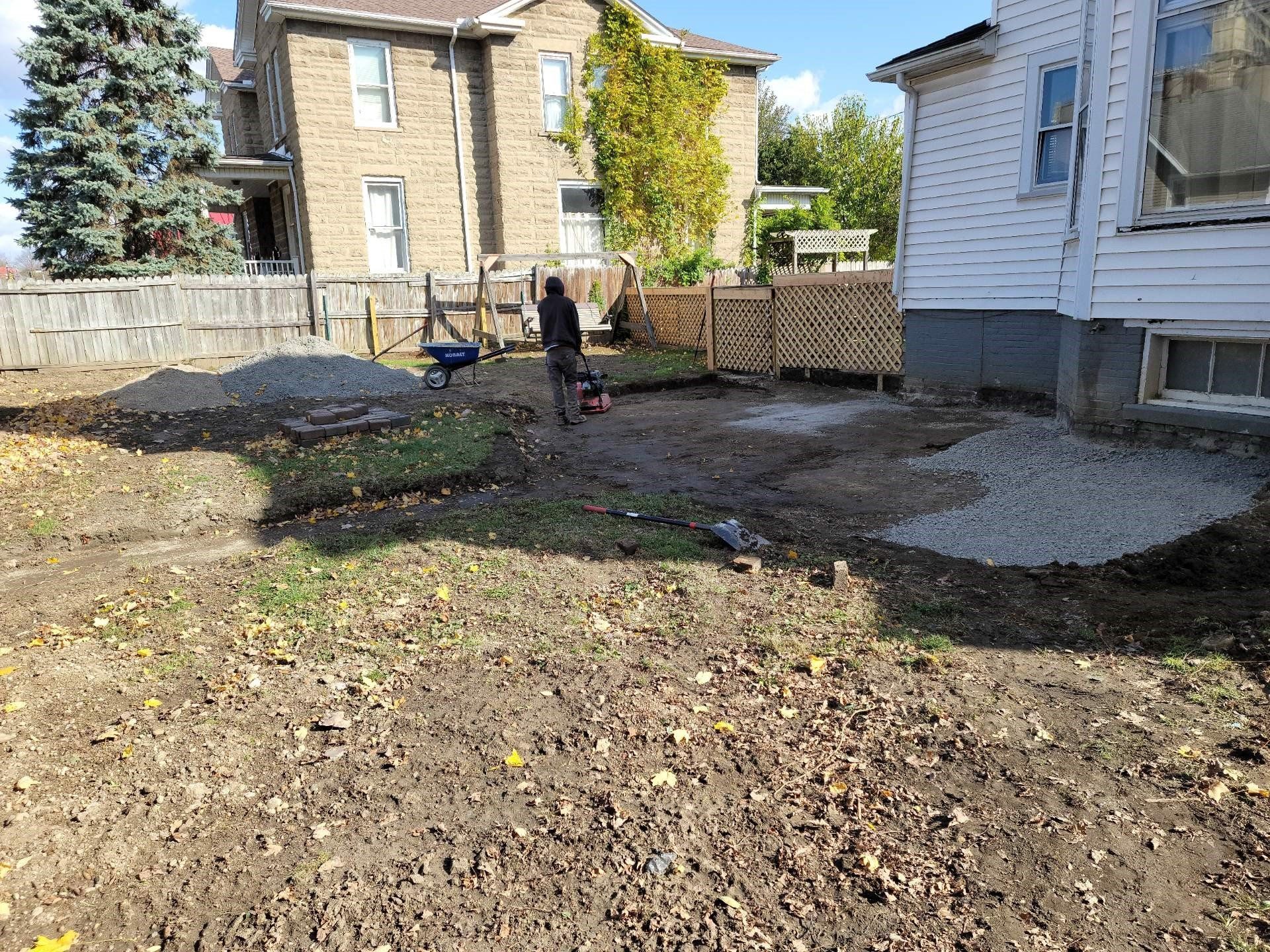 Backyard with a person working near a fence, rough bare ground, and houses in the background