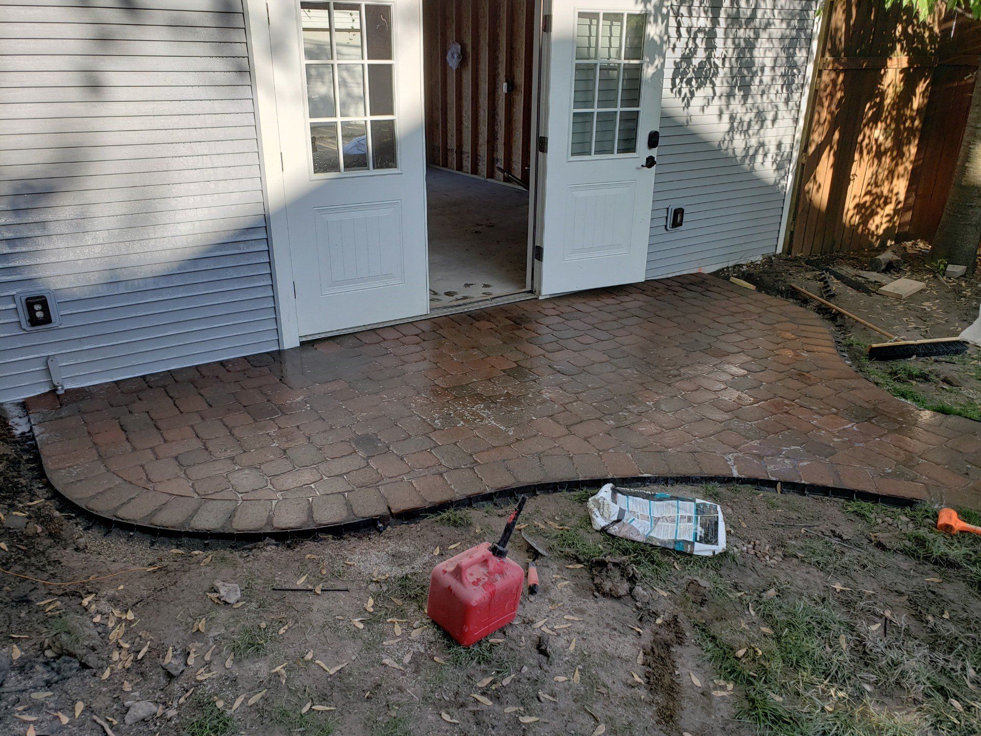 Brick patio with open white French doors, muddy ground, and a red gas can and bottle in the yard.