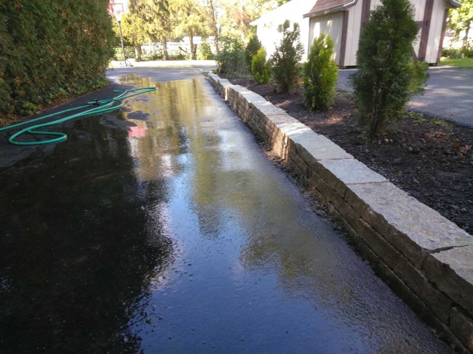 Flooded driveway with standing water reflecting trees and house, beside a garden hose and stone border