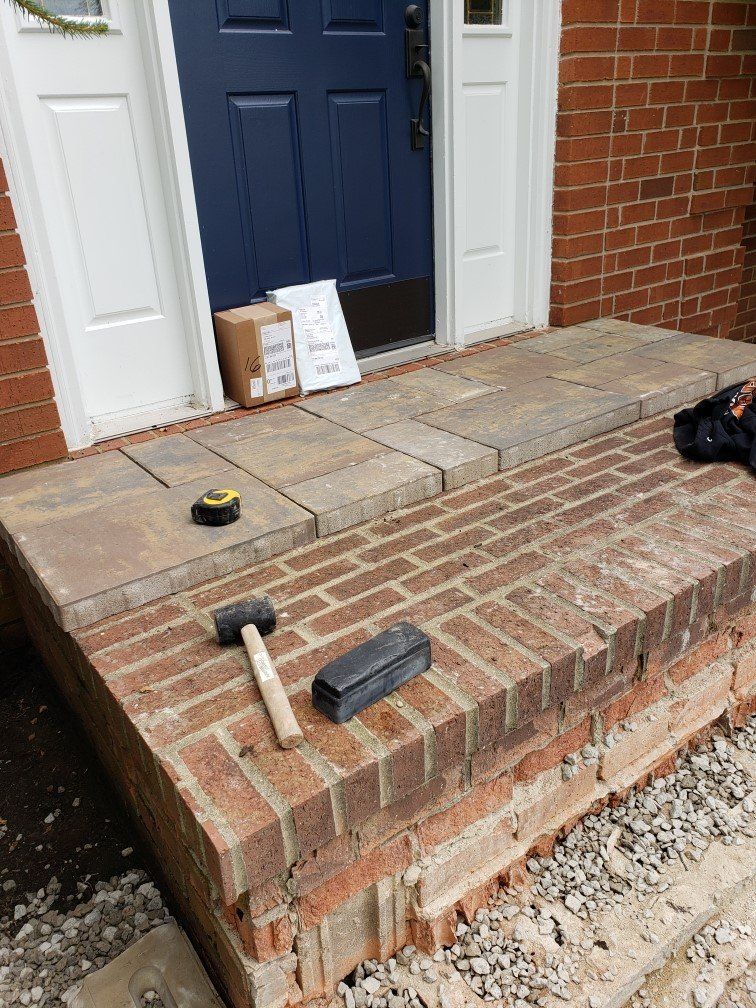 Front porch with tools and supplies near a blue door, showing brick steps and a partially unfinished walkway