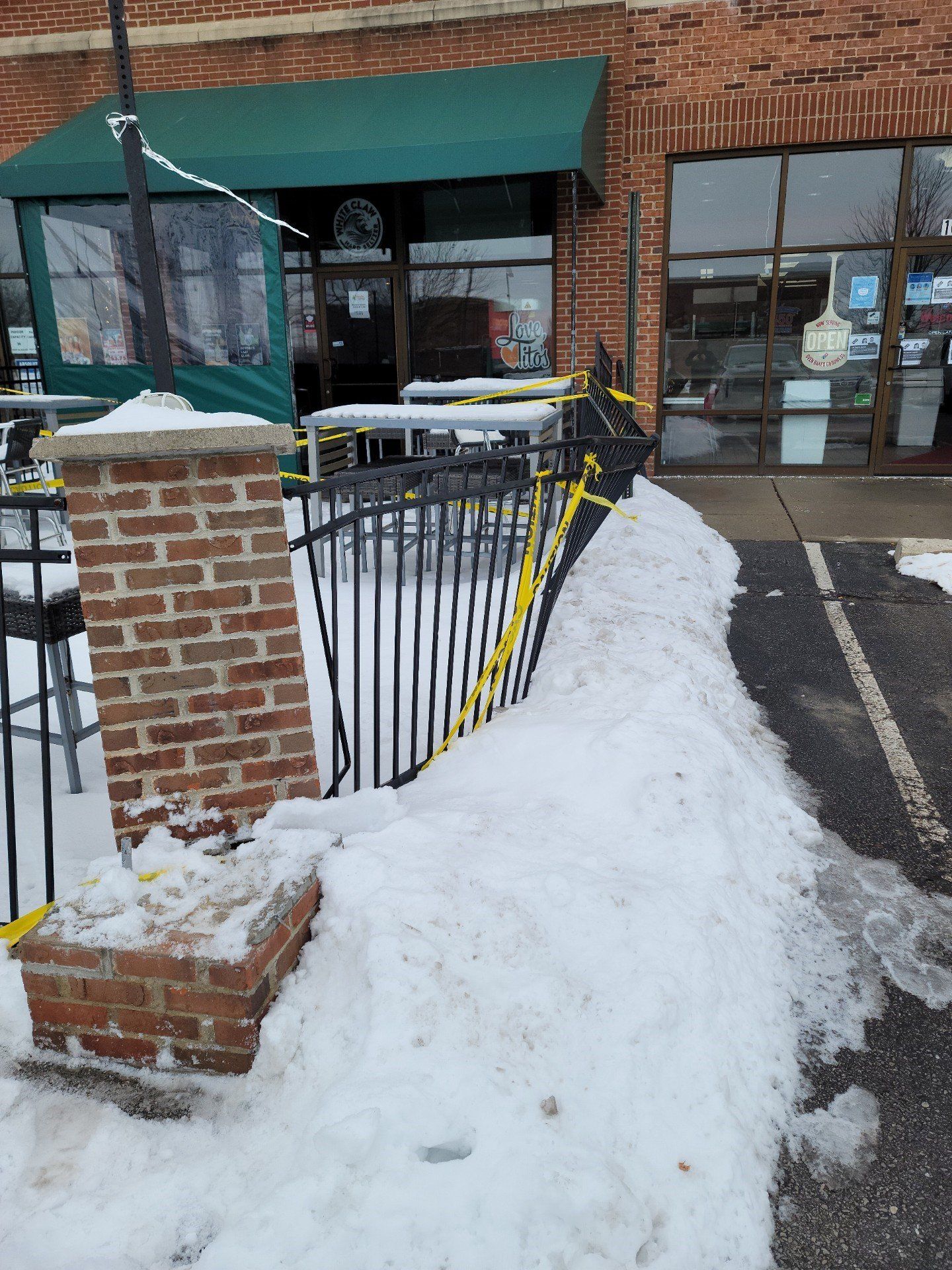 Snow-covered entrance with brick pillars, black railing, and green awning by a storefront
