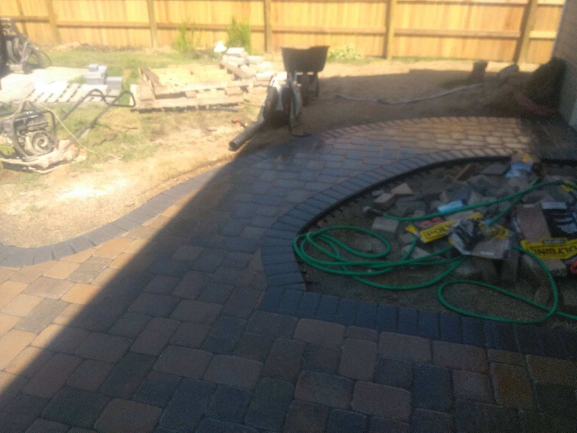 Backyard patio with brick pavers, coiled green hose, and a small black dog by the fence