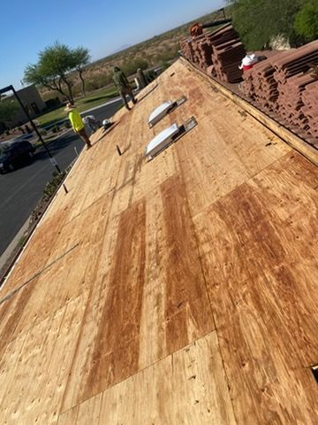 Roofer installing wooden roof sheathing on a sloped roof beside stacked red bricks and a worker in the background