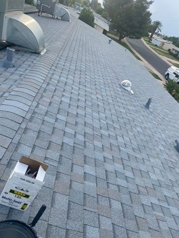 A high-angle view of a grey shingled roof with a ventilation box, plumbing vents, and an open box of roofing materials.