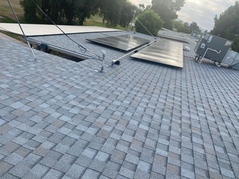 A gray shingled roof with solar panels installed, a metal vent pipe, and an HVAC unit under a cloudy sky.