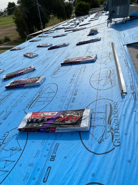 Bundles of roof shingles are spread across a residential roof covered in blue protective underlayment.
