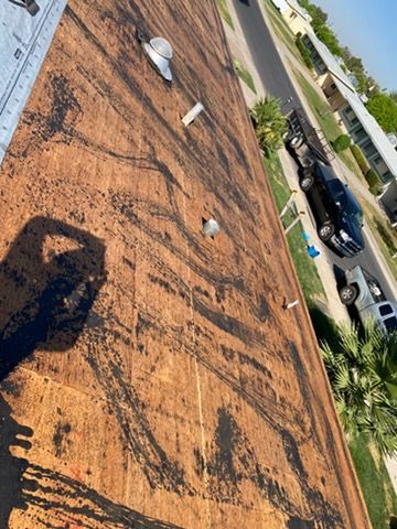 A view looking down from a rooftop, showing exposed plywood decking, vents, and a suburban street with vehicles below.