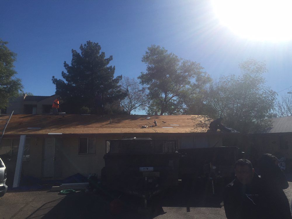 Workers on a roof removing old shingles under a bright sun. A dark truck and trees are in view.