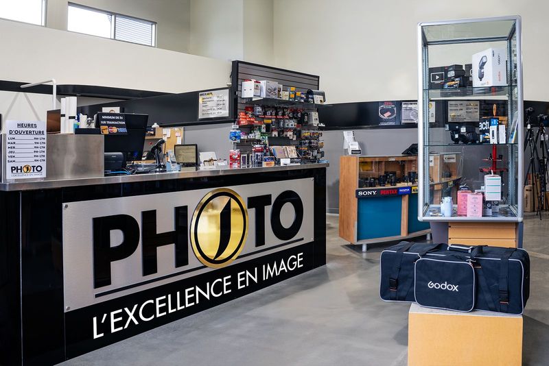Intérieur d'un magasin de photographie avec un comptoir et une vitrine. On peut lire sur le comptoir « PHOTO » et « L'EXCELLENCE EN IMAGE ».