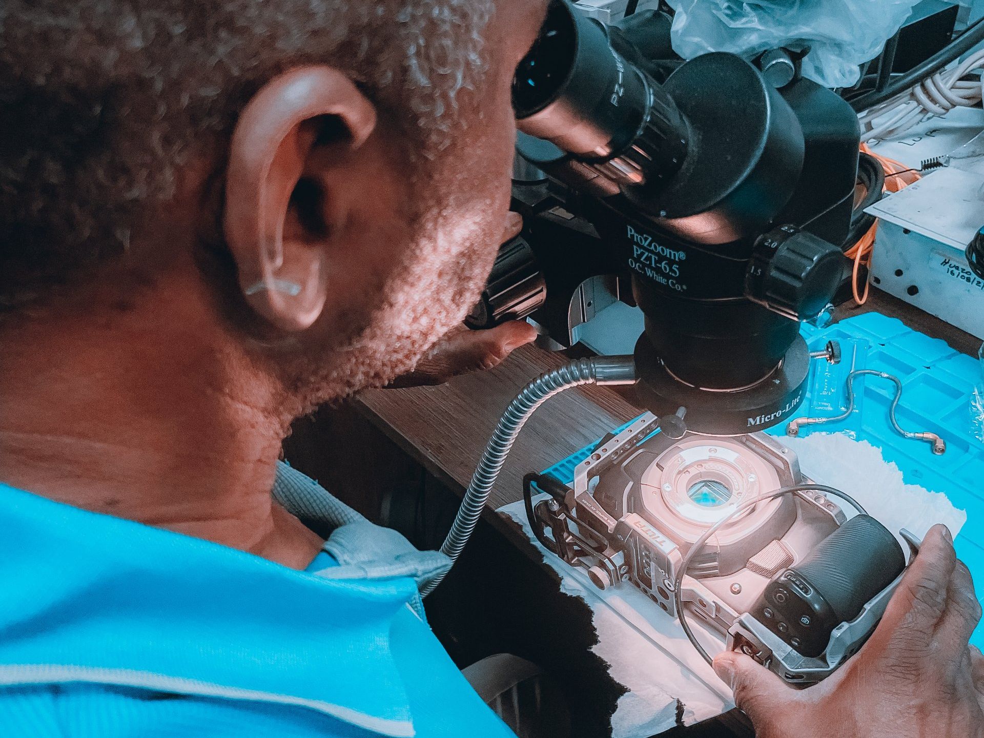 Un homme observe un petit appareil au microscope, probablement en train de le réparer. Éclairage bleu.