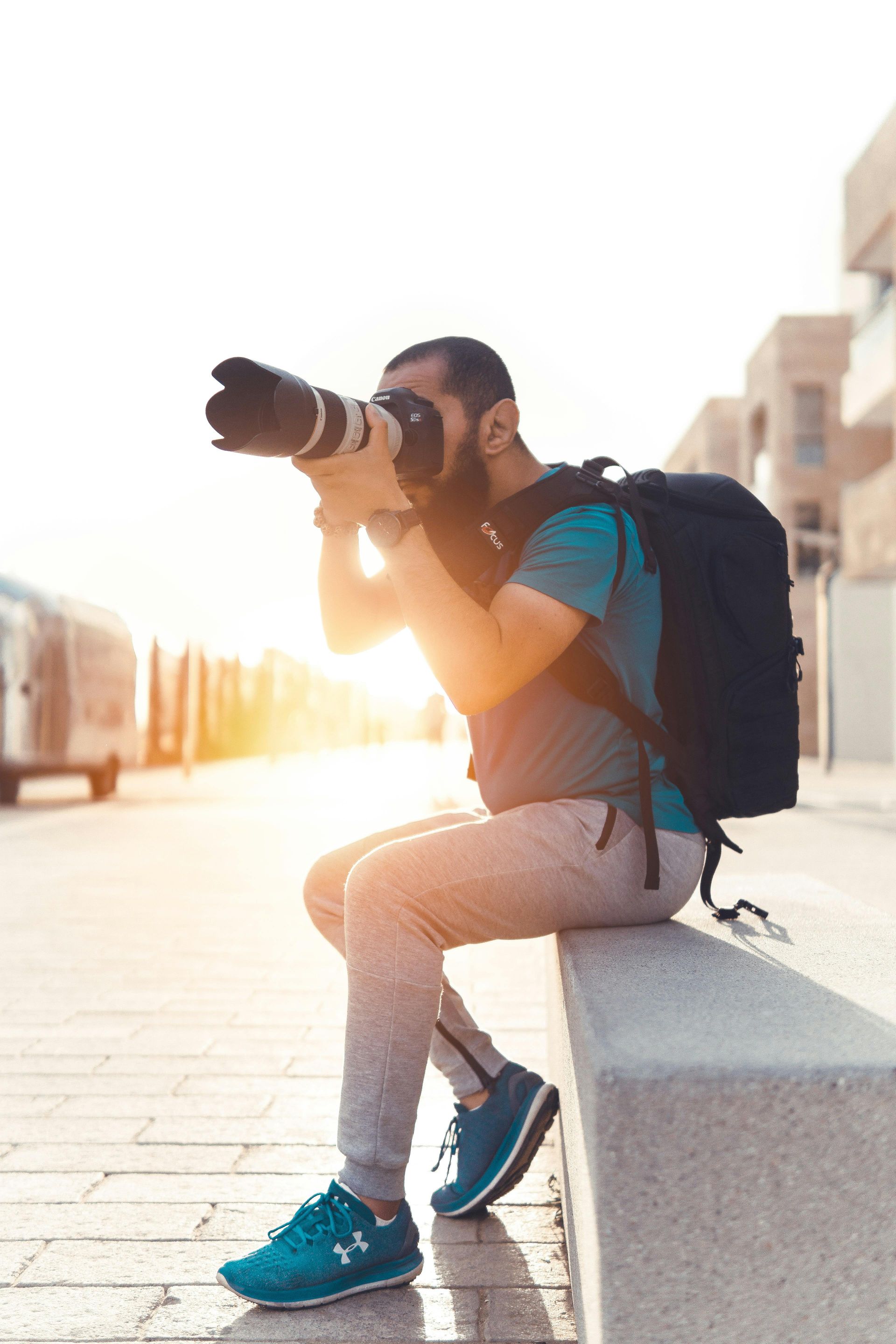 Un homme avec un appareil photo, assis dehors, prend une photo. Il porte un sac à dos et des baskets ; la lumière du soleil est en arrière-plan.