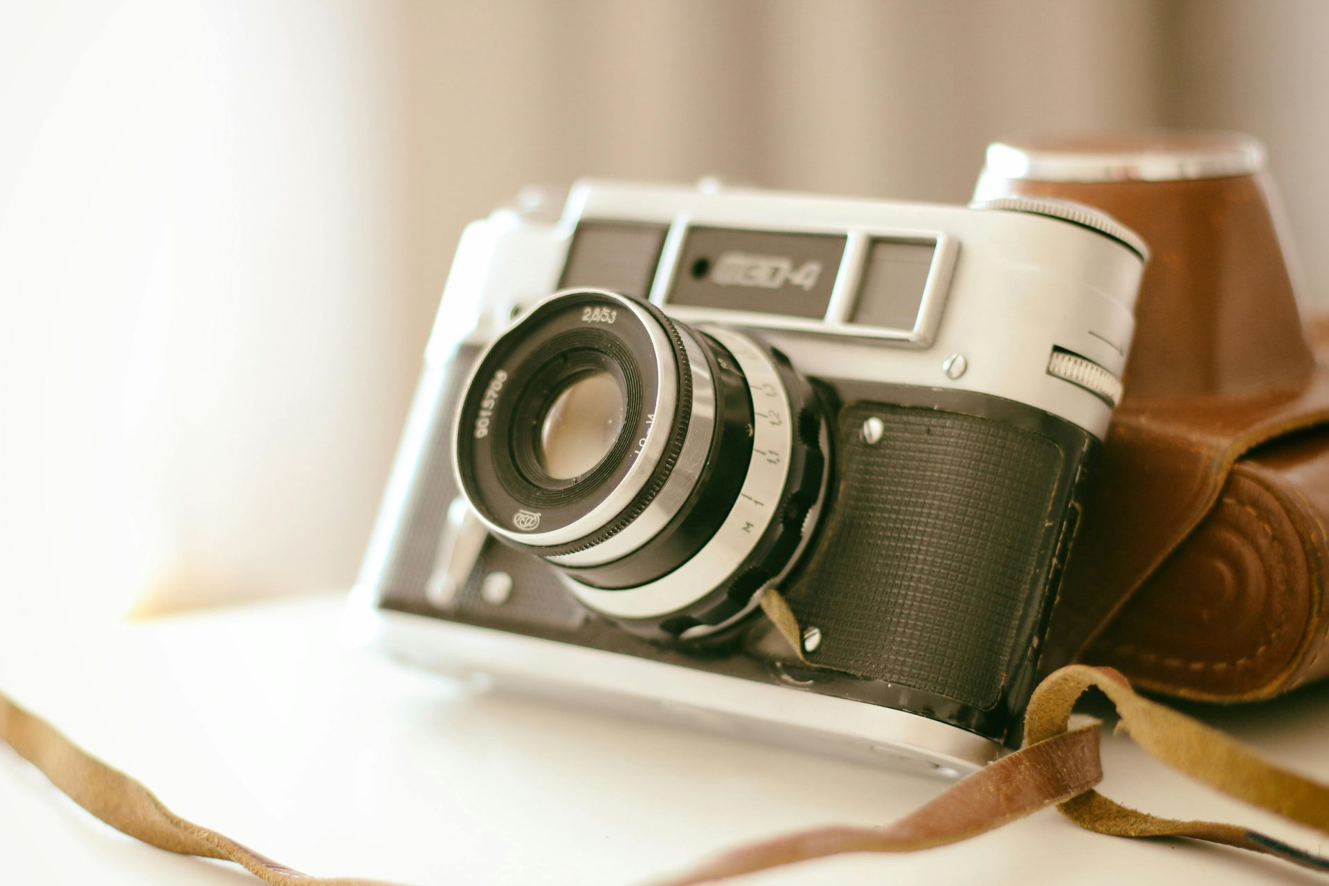 Appareil photo vintage argenté et noir avec étui en cuir marron, posé sur une surface blanche.