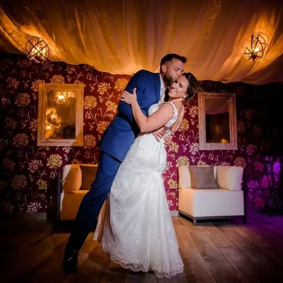 Groom dips bride, kisses her forehead; wedding scene with floral wallpaper and soft lighting.