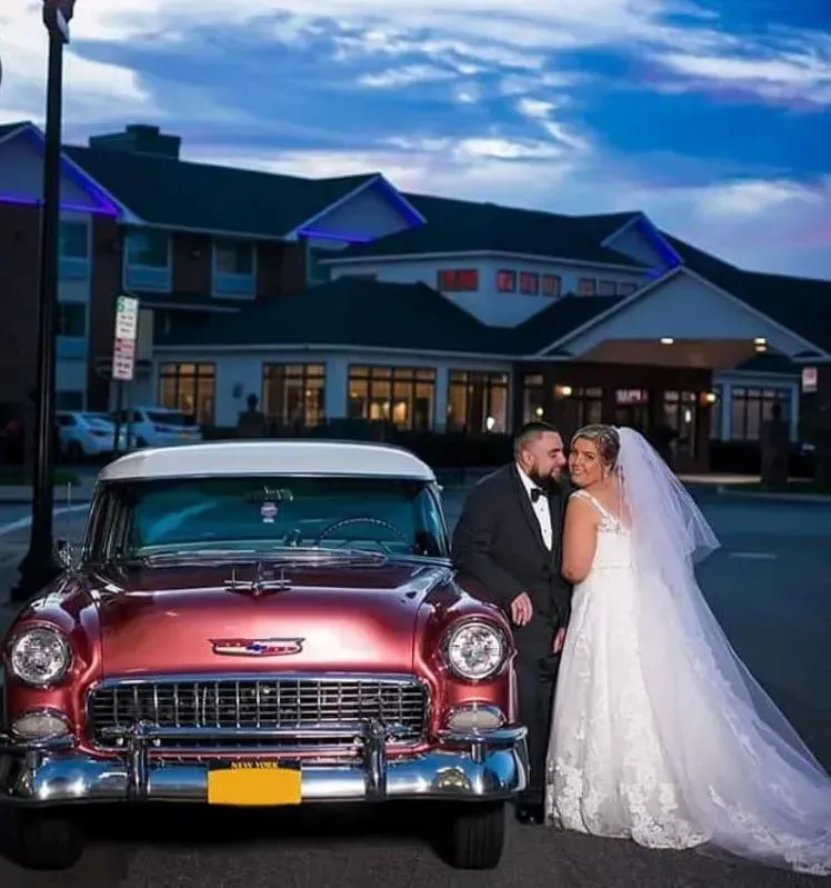 Newlyweds pose by a classic red car in front of a building at dusk. Bride in white dress, groom in black suit.