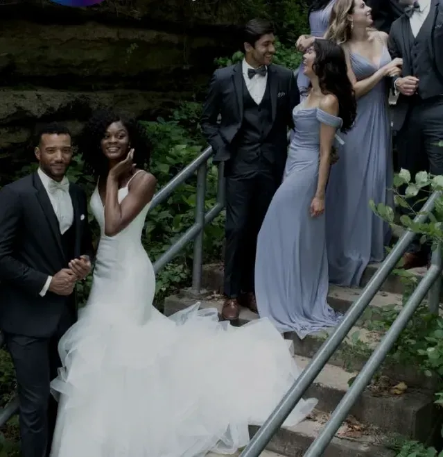 Wedding party on outdoor stairs: bride in white dress, groom in black suit, bridesmaids in blue.