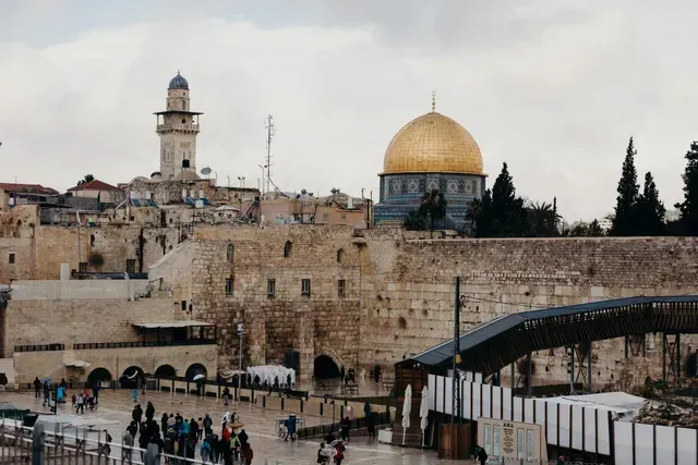 View of the Western Wall and Dome of the Rock in Jerusalem, with people in the plaza and cloudy sky.