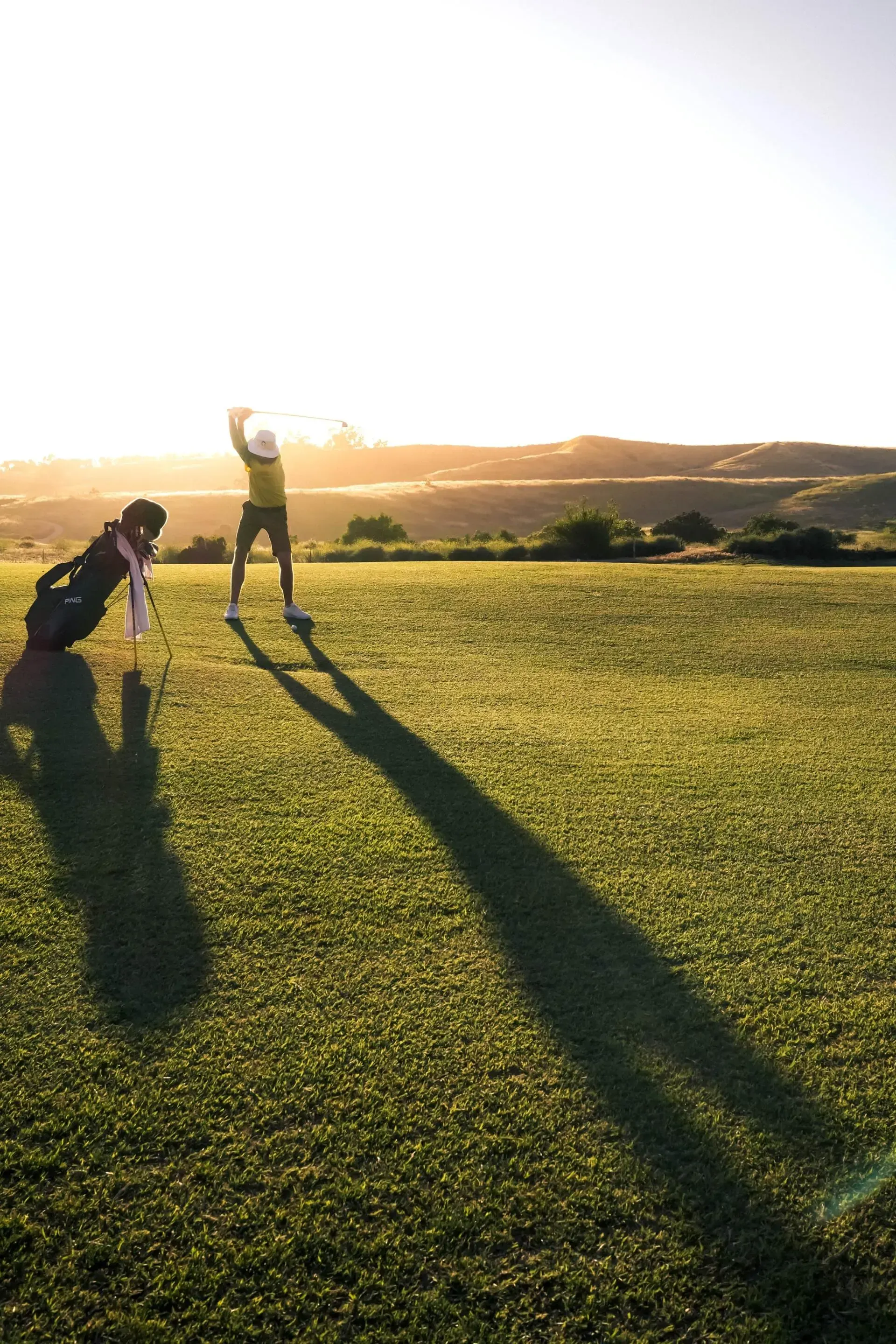 Golfer swings club on green, long shadow, other person tending golf bag, sunlit landscape.