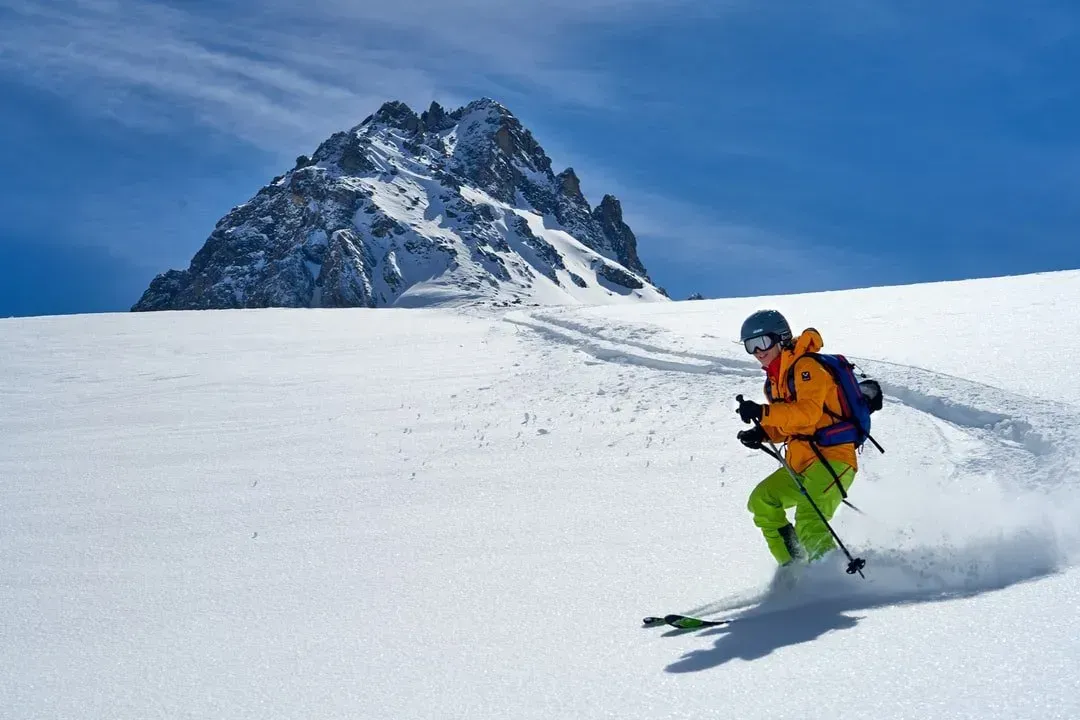 Skier in orange jacket and green pants skis down a snowy slope, mountain in background.
