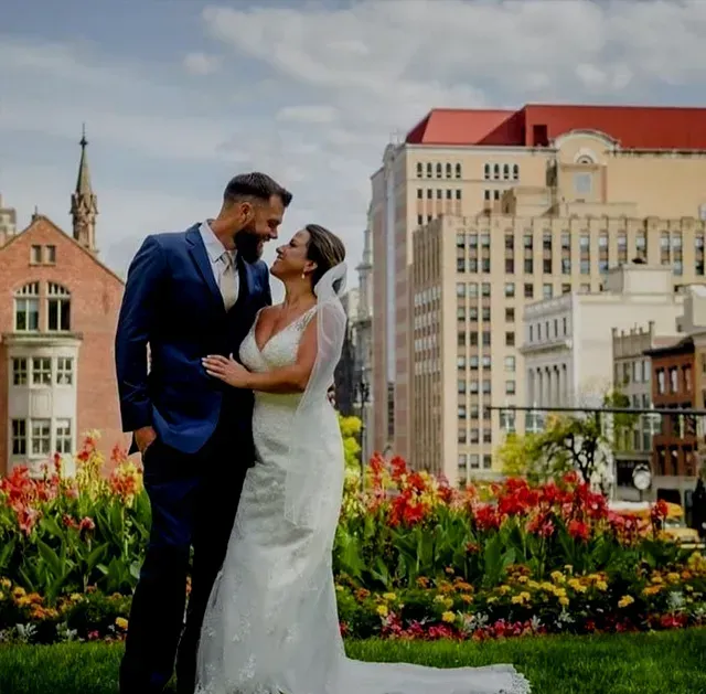 Bride and groom embracing in front of a city skyline, surrounded by colorful flowers.