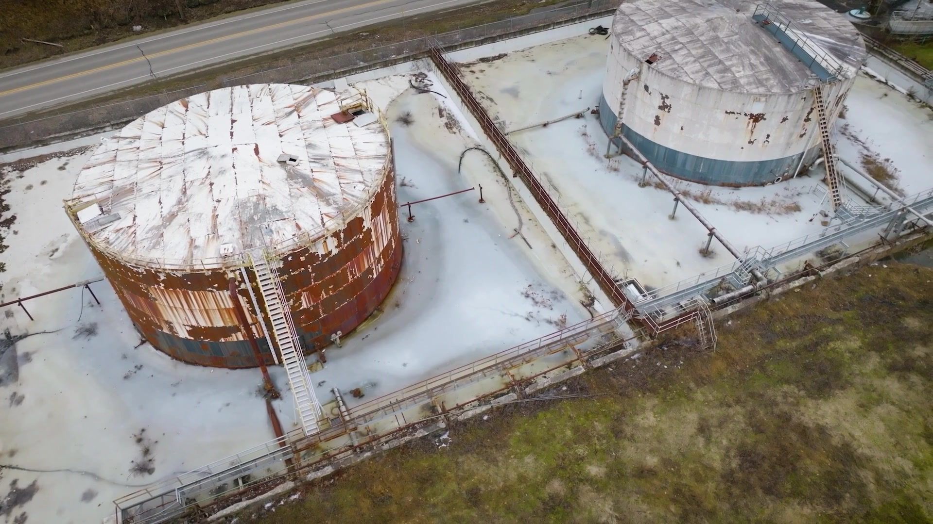 Aerial view of two large snow-covered cylindrical tanks beside a road and grassy field.