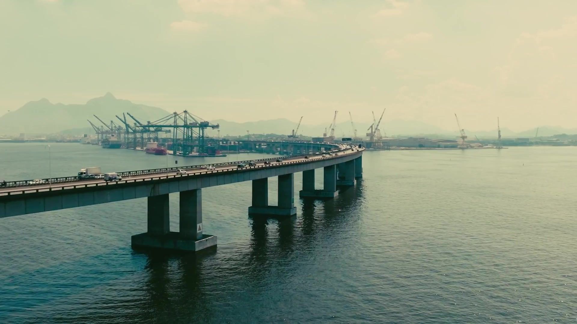 Long pier extending into calm water at a hazy industrial harbor with cranes in the distance
