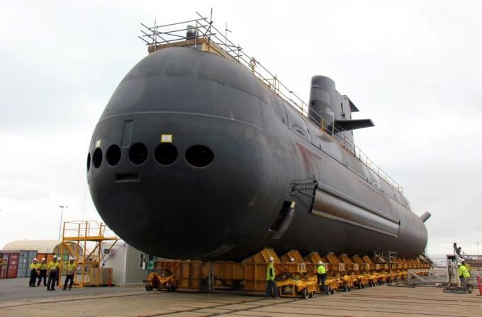 Large black submarine under construction on a dock, with workers and scaffolding around it.