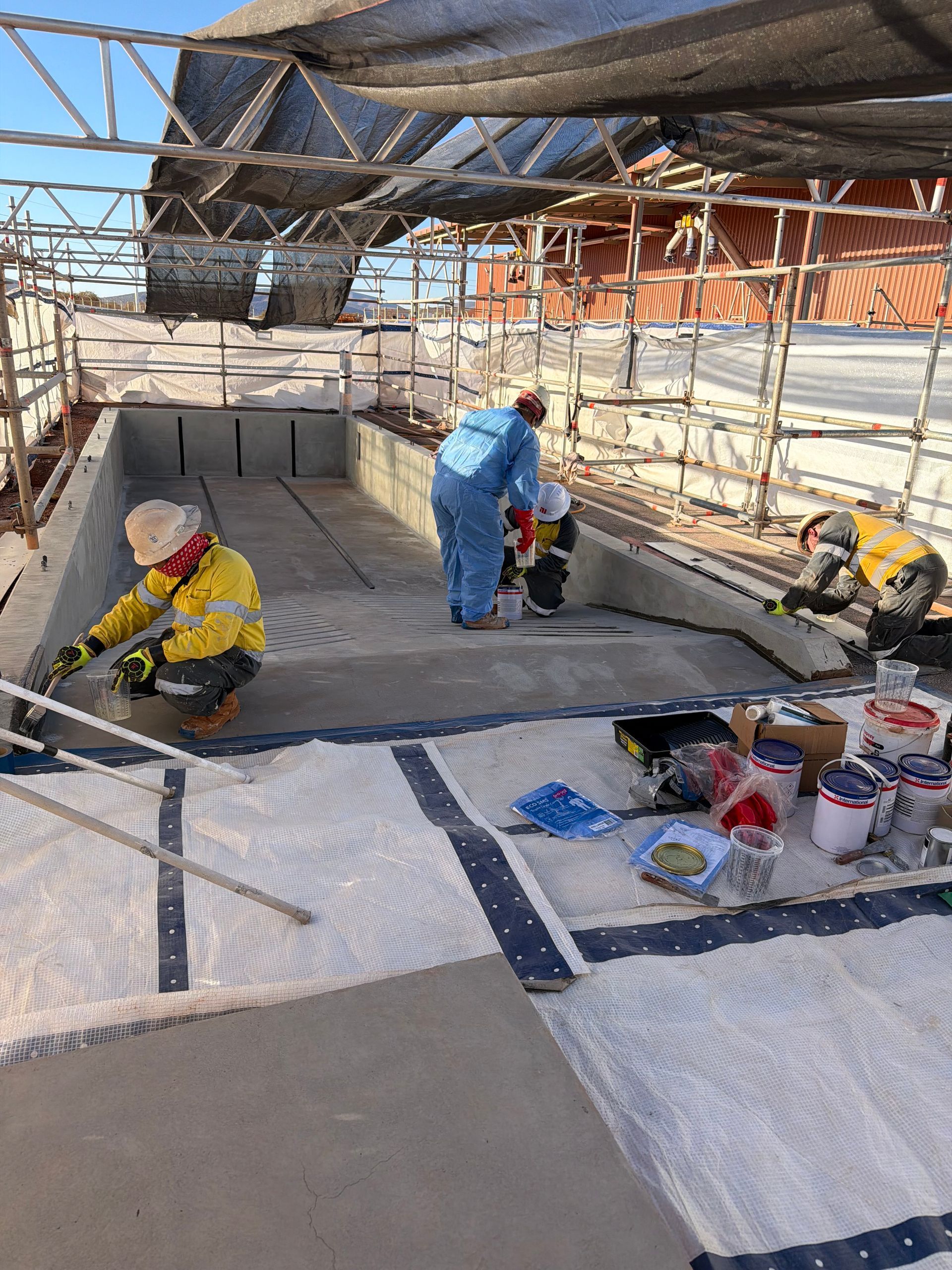 Workers in blue and yellow gear sanding a floor in an unfinished building with scaffolding