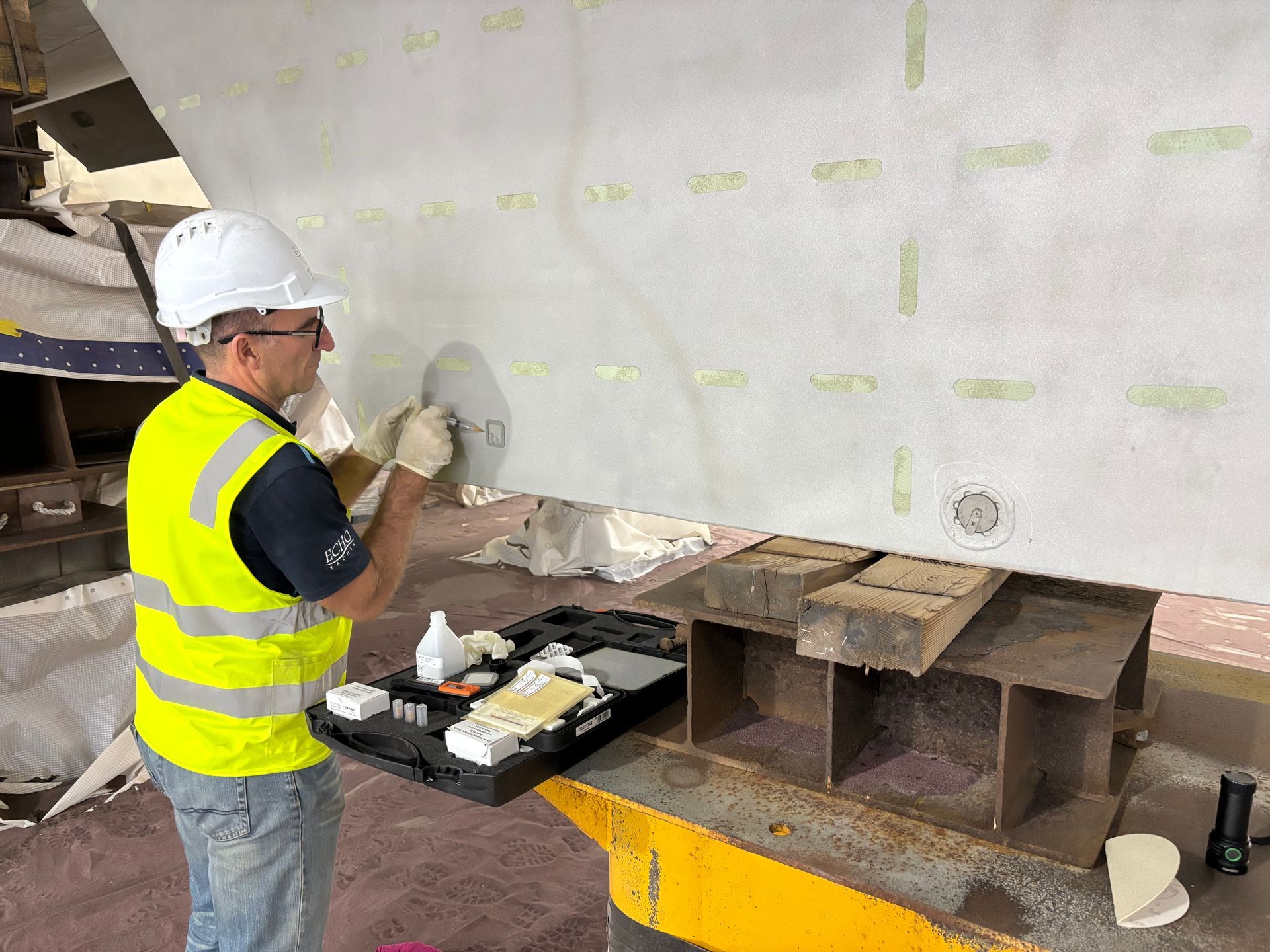 Worker in a hard hat and hi-vis vest applying material to a large metal surface at a job site.