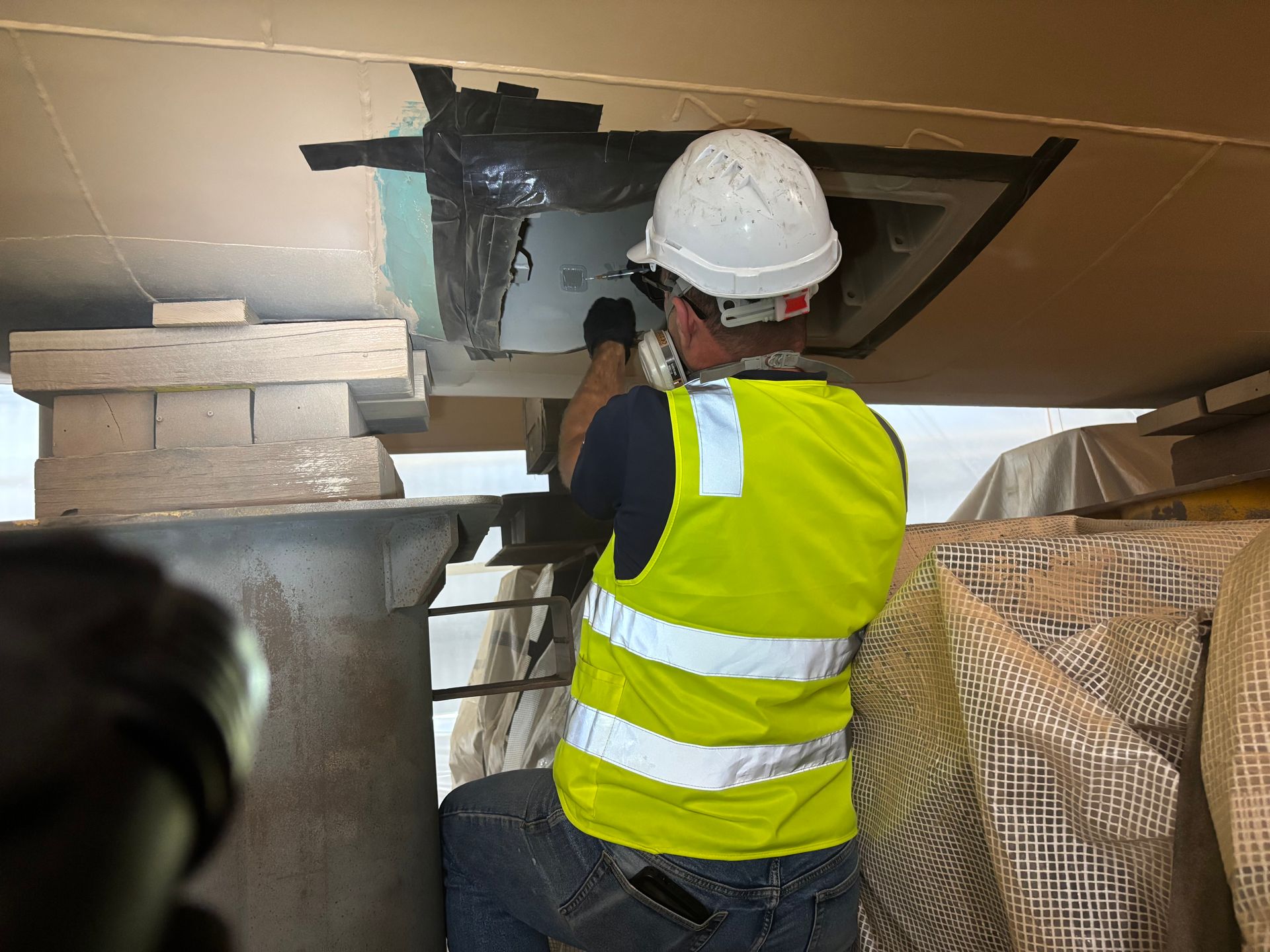 Worker in hard hat and hi-vis vest inspecting a damaged ceiling opening in an indoor construction site.
