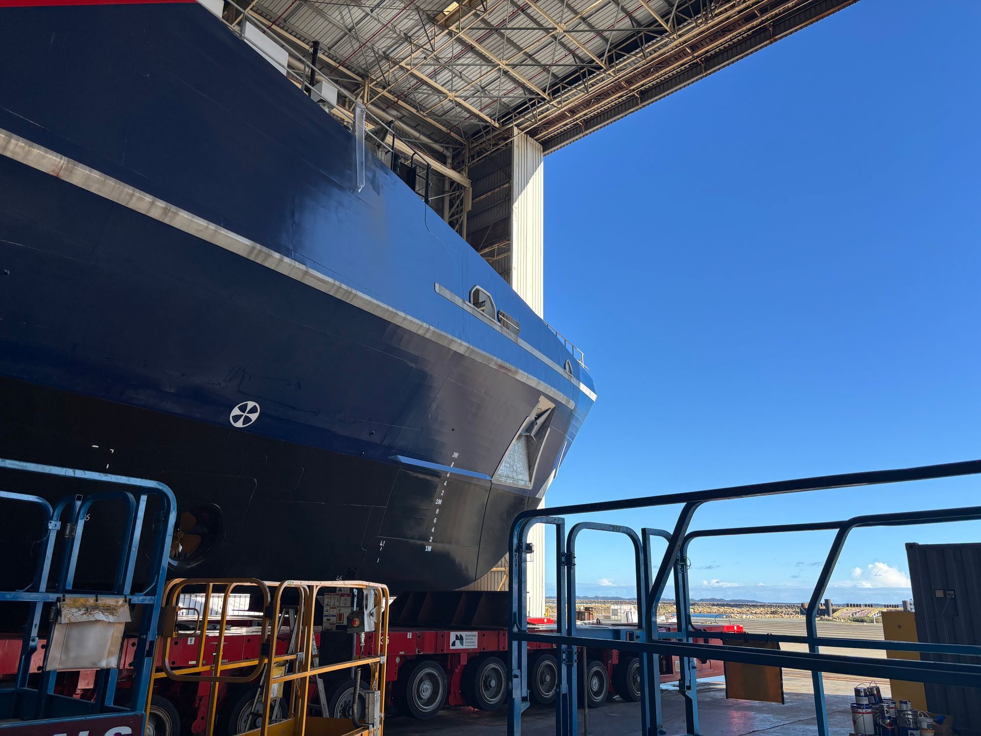 Large blue ship in dry dock under a roof, with support equipment and railings in the foreground.